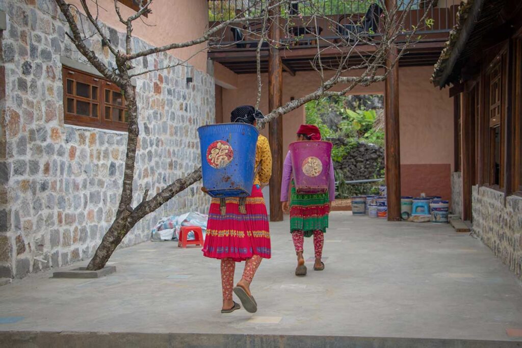 Local Hmong women in traditional clothing carrying baskets through the courtyard of the nearly finished Ha Giang mountain lodge, showing real community involvement in construction.