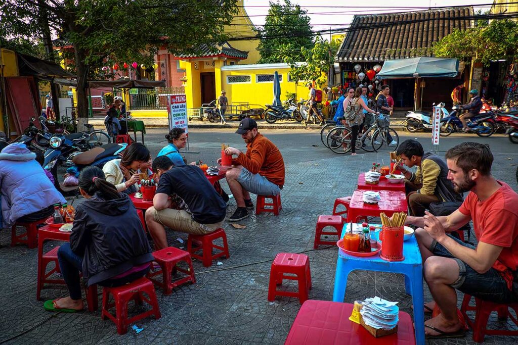 Backpackers in Vietnam are eating street food at a local food stall in Hoi An while sitting on low plastic stools on the street