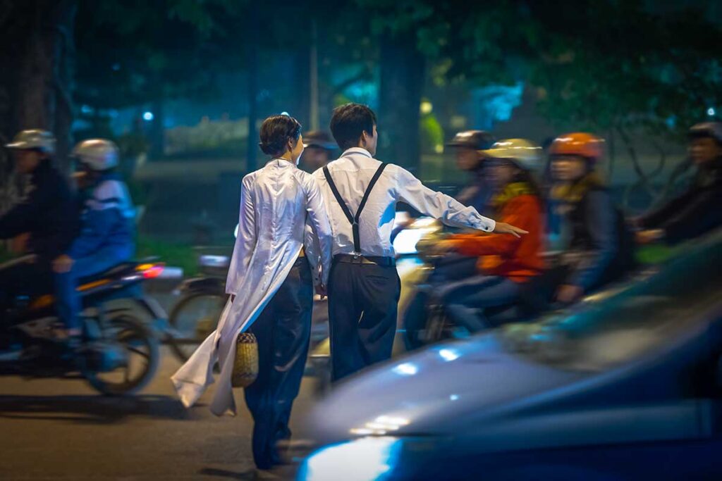 a man and a woman are crossing a road in Vietnam through busy traffic of motorbikes and cars