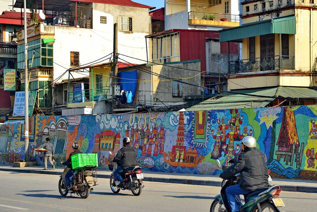 Hanoi Ceramic Mosaic Mural Street Scene: Motorbikes zip past a vibrant section of the Hanoi Ceramic Mosaic Mural, depicting daily life in Vietnam.