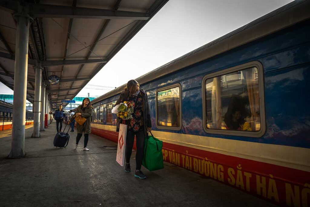 Vietnamese travelers carrying luggage on the platform beside a parked train at Hanoi Railway Station.