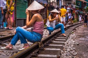 A young woman captures a photo of her friend wearing a traditional Vietnamese conical hat on Hanoi's iconic Train Street, a popular Instagram spot.