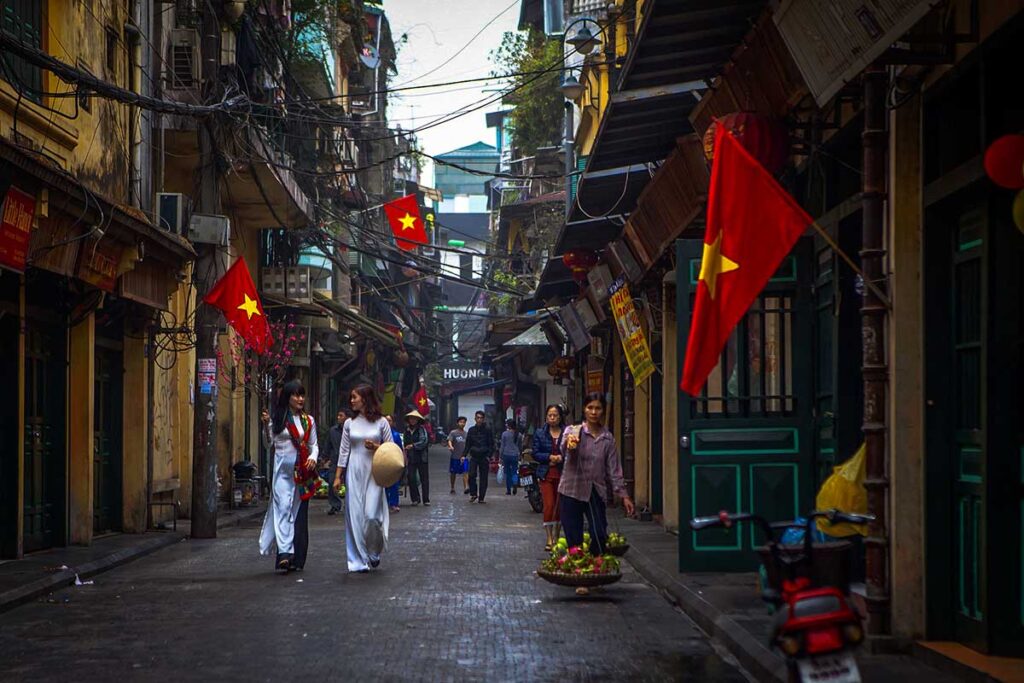 Ta Hien Street, Hanoi Old Quarter: A street vendor carries a traditional shoulder pole laden with baskets of fruit on Ta Hien Street in Hanoi's Old Quarter. Vietnamese women in white Ao Dai stroll past colorful shops adorned with Vietnamese flags.