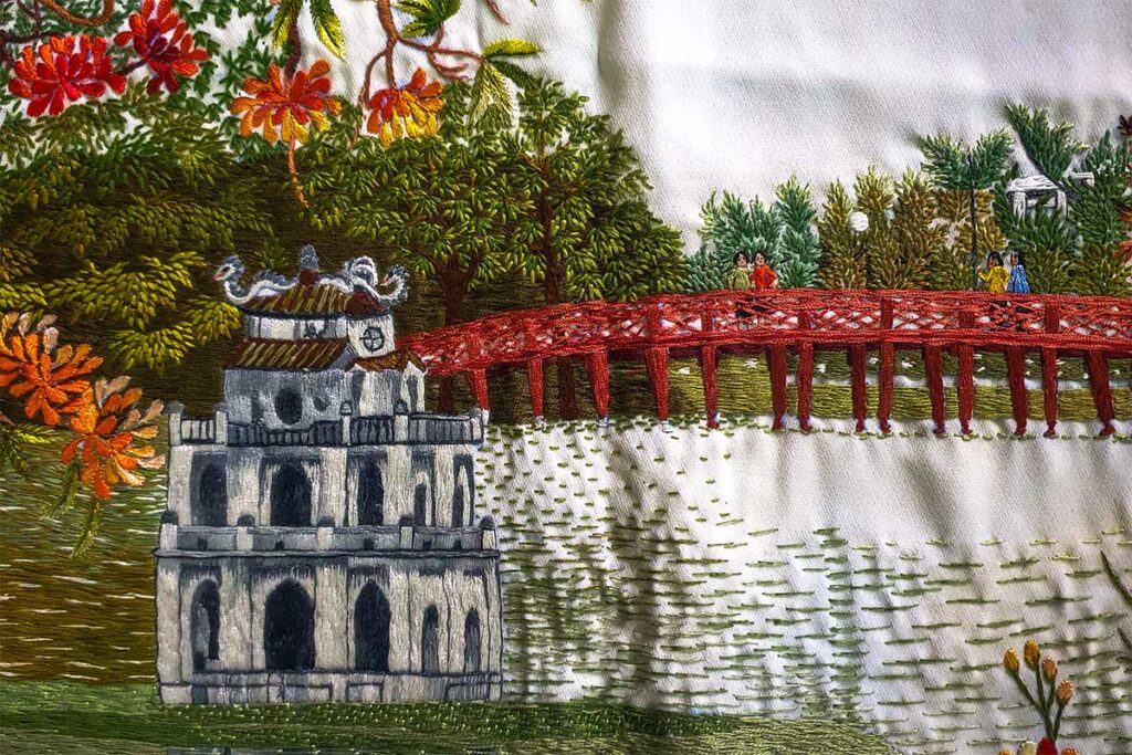 A detailed embroidered rug depicts Hanoi's iconic Hoan Kiem Lake and Turtle Tower, a masterpiece from Quat Dong Village.