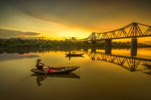 Long Bien Bridge at Sunset on Red River: The iconic Long Bien Bridge silhouetted against a golden sunset over the Red River, with rowing boats dotting the water.