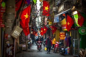 A bustling street scene in Hanoi, a vibrant city in Vietnam, with motorbikes zipping through narrow lanes, locals walking past colorful shophouses adorned with Vietnamese flags.