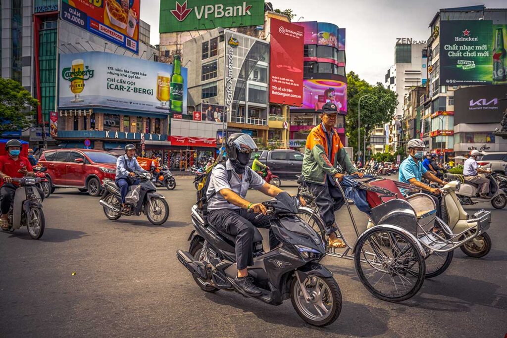 Typical Vietnamese street scene featuring a cyclo, surrounded by motorcycles and a few cars on a busy street.