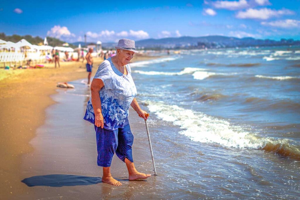 Elderly woman with a cane standing on a sandy beach, representing accessible travel and enjoying vacations despite mobility challenges.