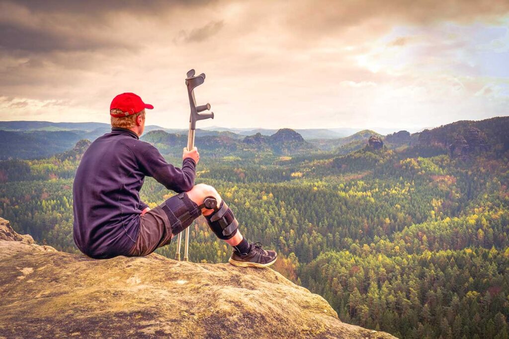 Man with crutches sitting on a hilltop after a hike, overlooking a stunning scenic view, illustrating travel with disabilities