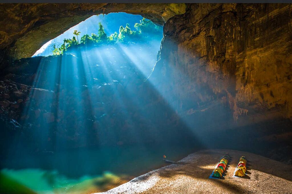 Light beams coming through a cave entrance shining on a campsite next to an underground lake at Hang En Cave in Phong Nha