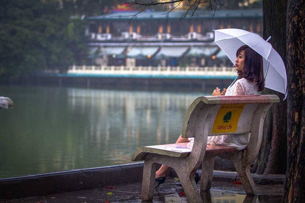 A woman sitting on a bench while holding an umbrella and watching Hoan Kiem Lake during the rain in Hanoi