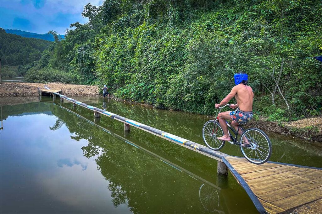 A man cycling over a narrow beam over a pond - A game called Monkey Bridge at Cuong Rung Farm in Phong Nha