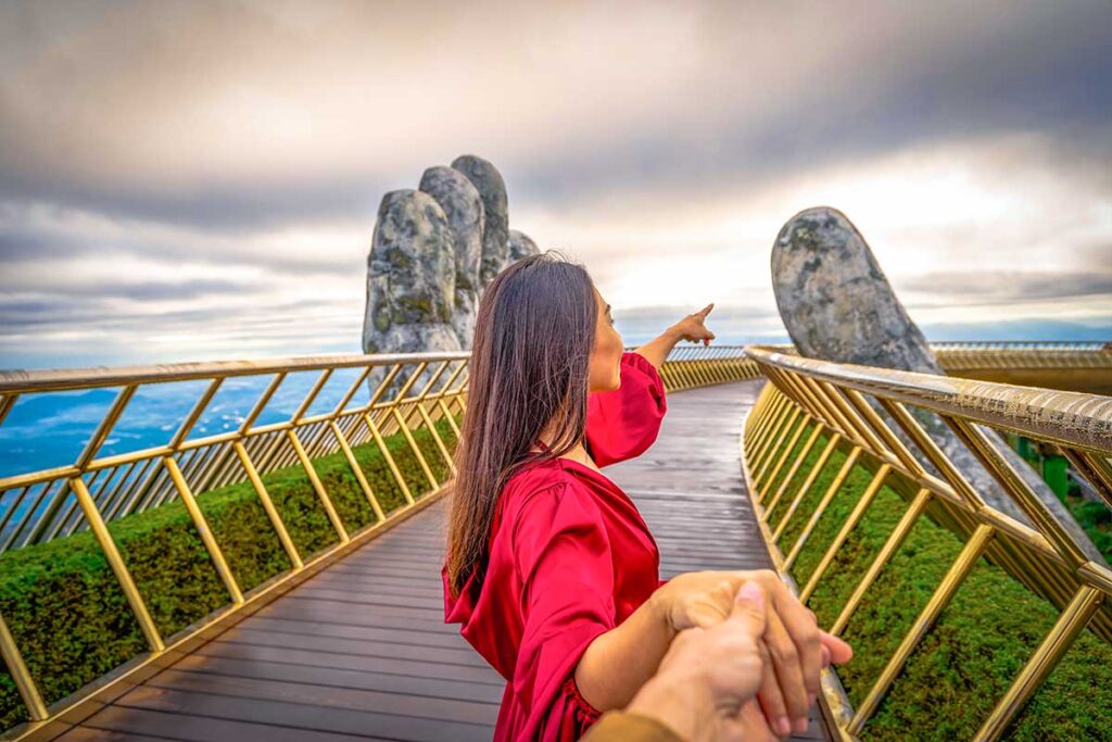 A romantic view of a woman with a red dress holding the hand of her partner on the Golden Hand Bridge at Ba Na Hills in Da Nang