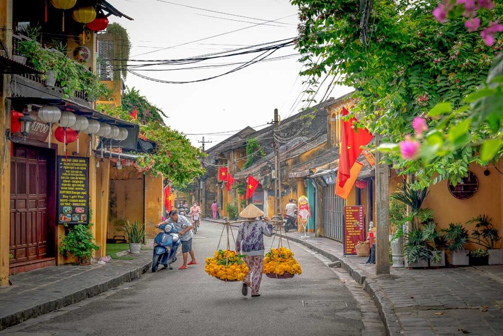 Local vendor carrying baskets of flowers through the quiet streets of Hoi An Ancient Town – a timeless daily scene in Vietnam.