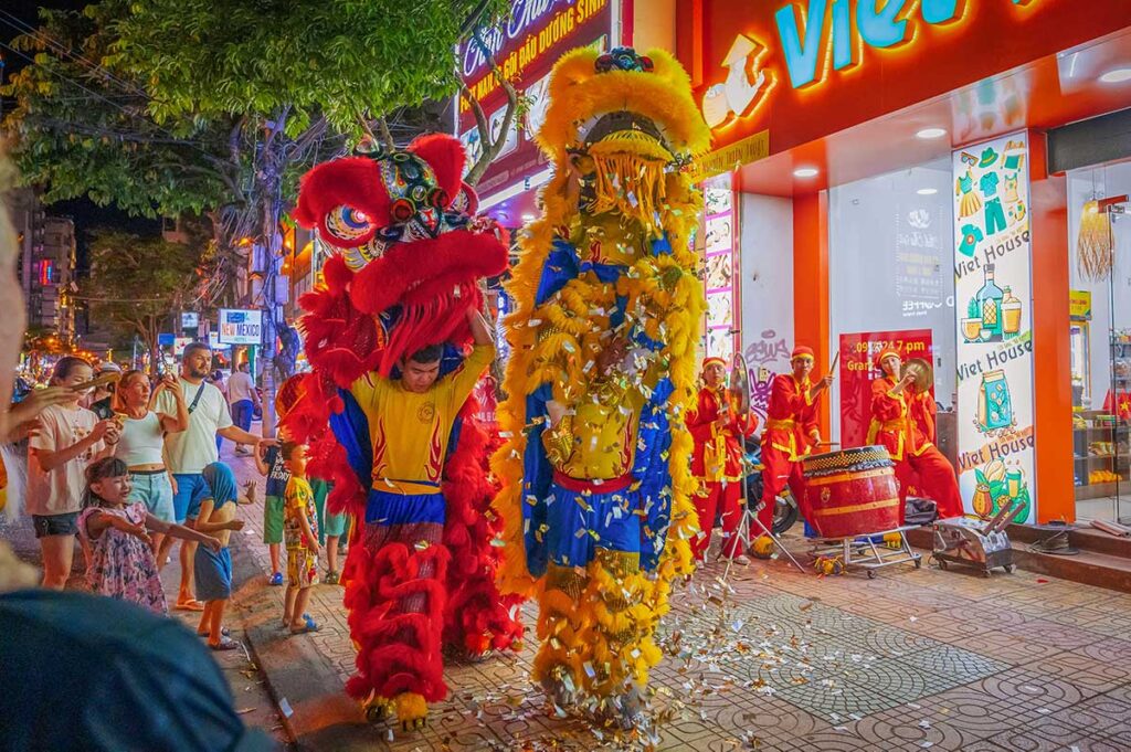 Vietnamese lion dance performance in front of a shop during a grand opening ceremony, with drummers and performers on the sidewalk.
