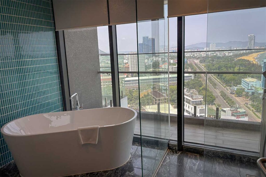 Modern Quy Nhon hotel bathroom with panoramic glass windows overlooking the city skyline and coastline, offering luxury sea-view stays.