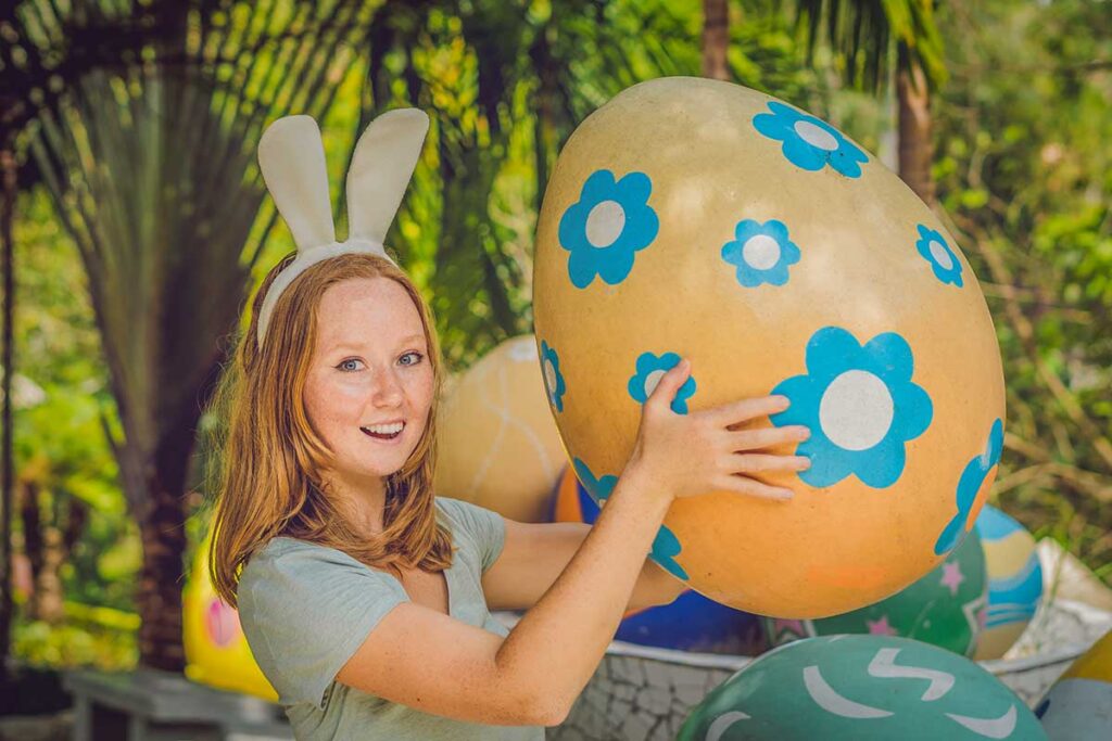 Tourist celebrating Easter in Vietnam surrounded by colorful Easter egg decorations in a tropical garden setting.