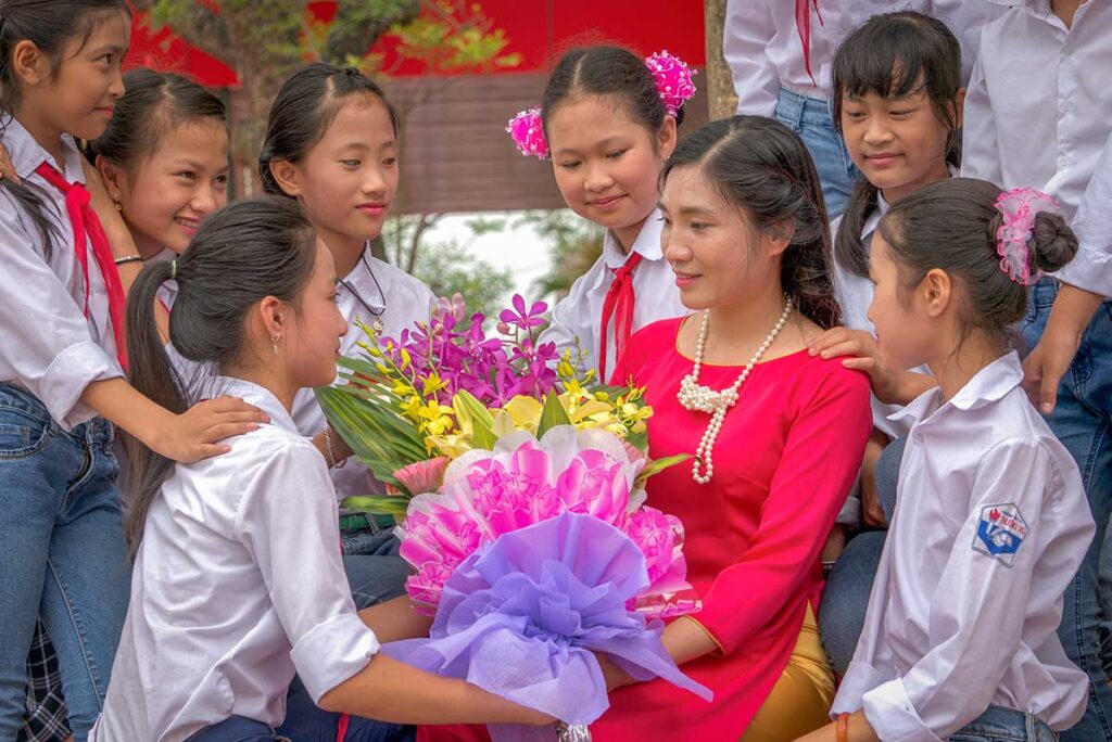 Vietnamese teacher surrounded by students holding flowers as part of Vietnamese Teachers’ Day traditions.