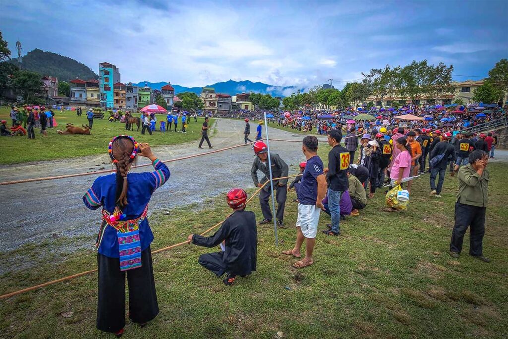 Group of ethnic people sitting and standing along the track of a horse racing event in Bac Ha