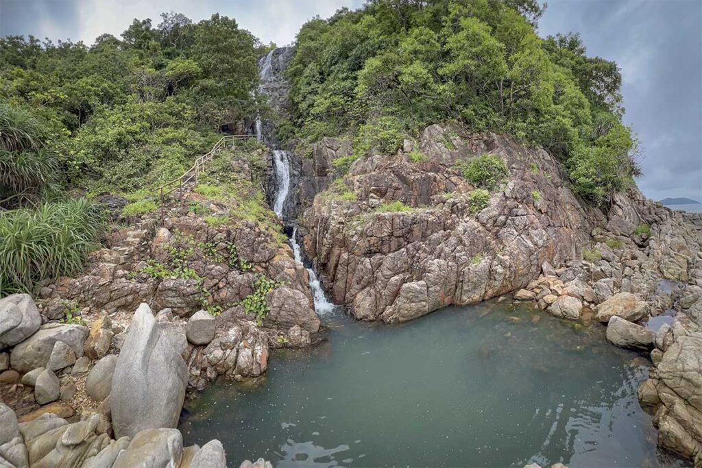 Small waterfall flowing into a natural pool along the Bai Bang hiking trail in Con Dao National Park, surrounded by rugged rocks and dense green vegetation