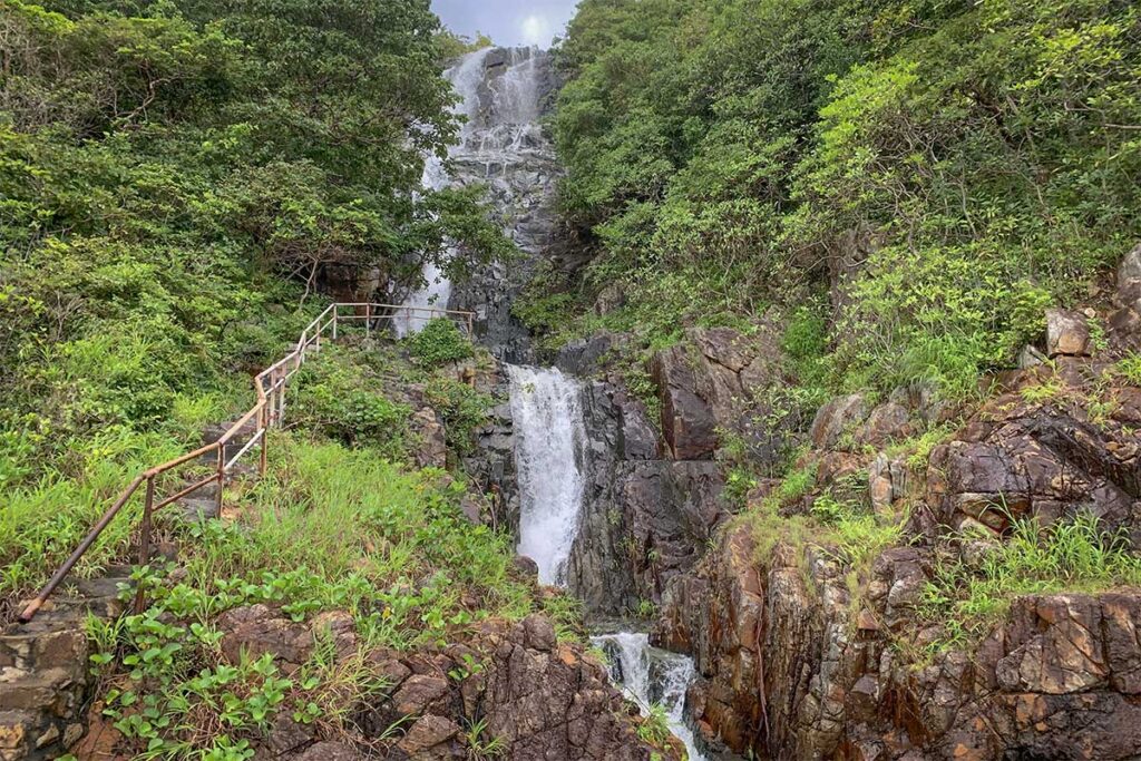 Waterfall cascading down rocky cliffs along the Bai Bang jungle trail in Con Dao National Park, surrounded by dense tropical greenery and a narrow hiking path with railings