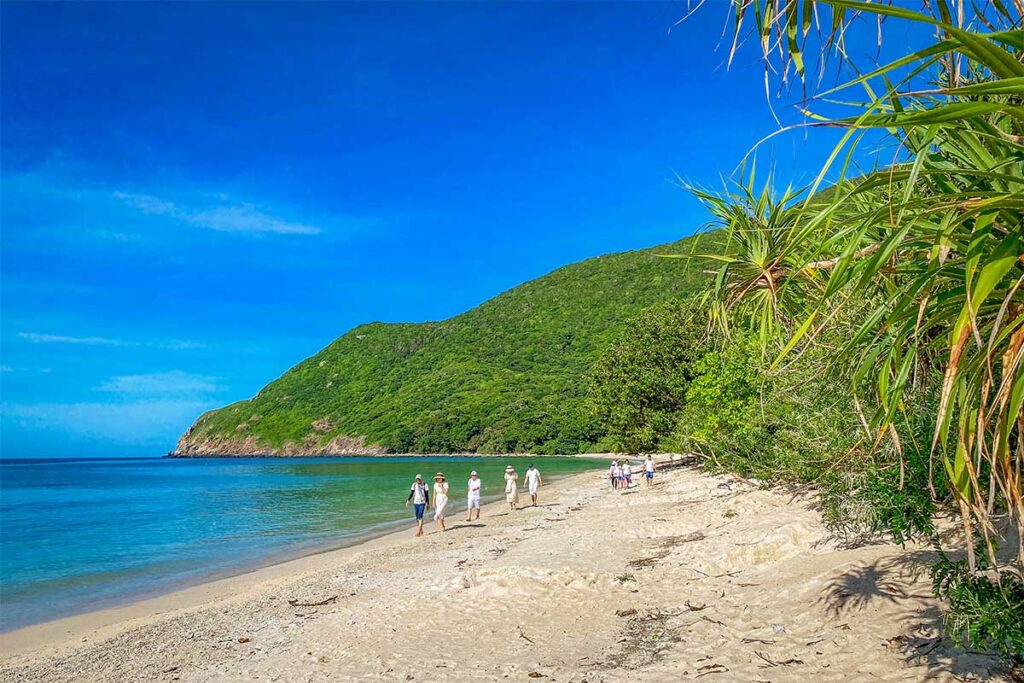 Quiet beach on Bay Can Island in Con Dao with clear turquoise water and a small group of visitors walking along the shoreline surrounded by green hills.