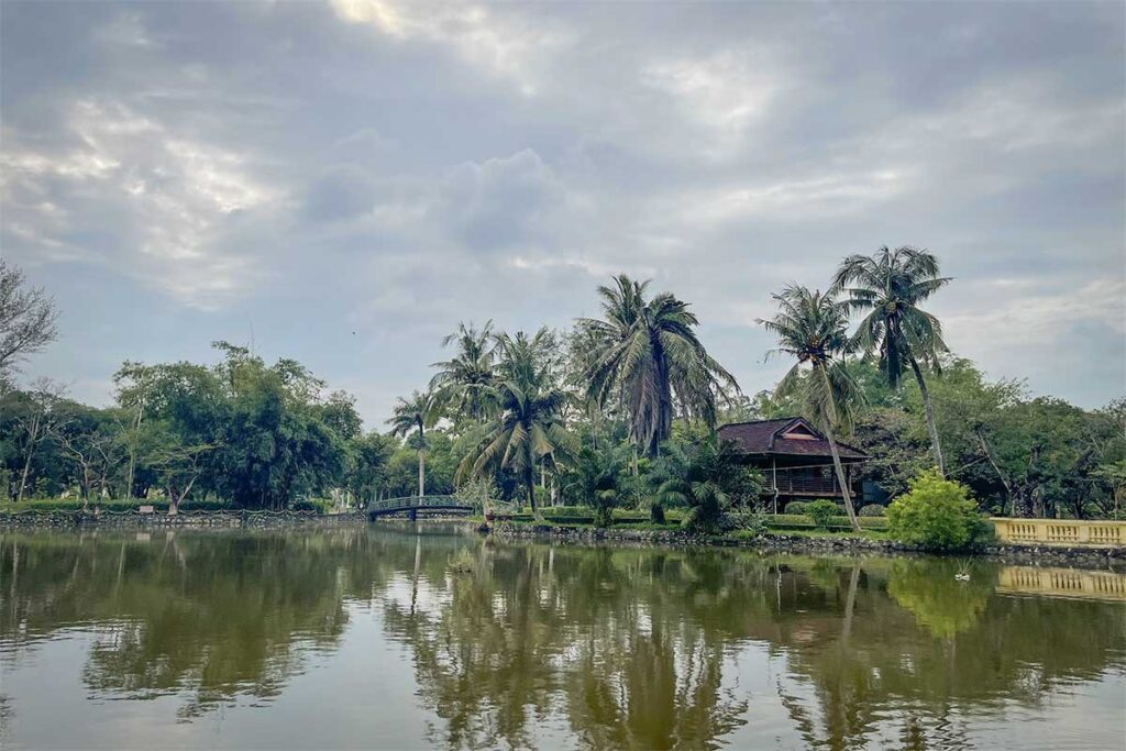 Scenic view of Ca Mau City Bird Garden with palm trees, wooden houses, and a lake surrounded by greenery.