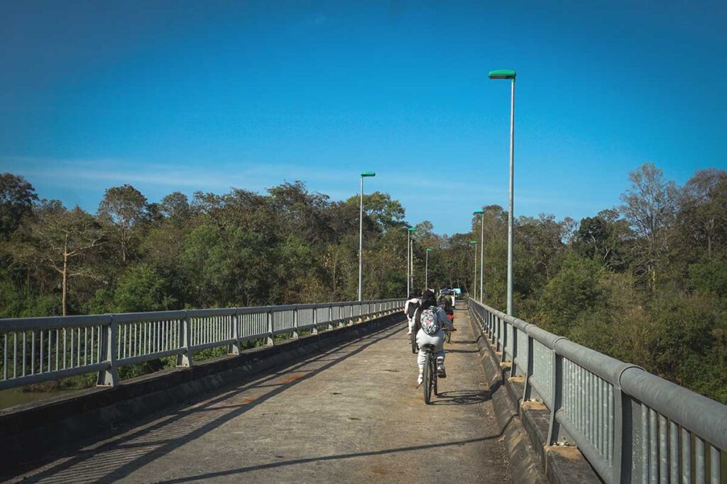 A group of tourists riding bicycles across a bridge, entering the lush surroundings of Yok Don National Park.