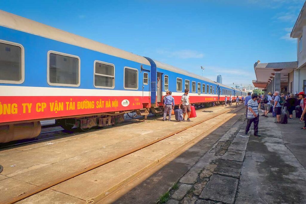 Travelers walking along the platform at a Vietnam train station, carrying luggage next to a stopped Reunification Express train