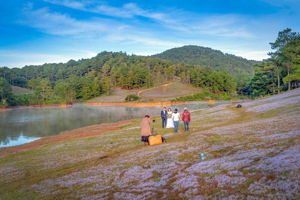 Couple taking wedding photos by a lake in Da Lat, with a photographer capturing the scene among pine-covered hills