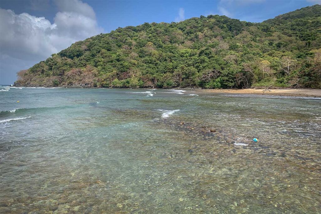Shallow clear water at Dat Tham Beach in Con Dao with small waves and a quiet coastline backed by dense green forest.