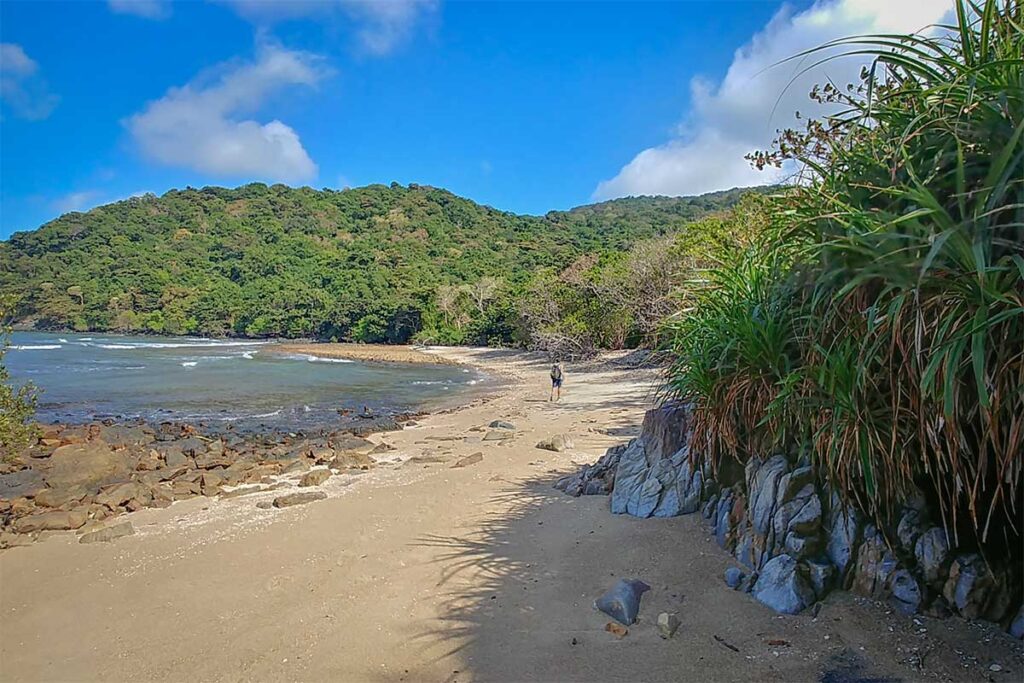 Quiet sandy shoreline at Dat Tham Beach in Con Dao National Park, with a lone traveler walking along the remote coast backed by forested hills
