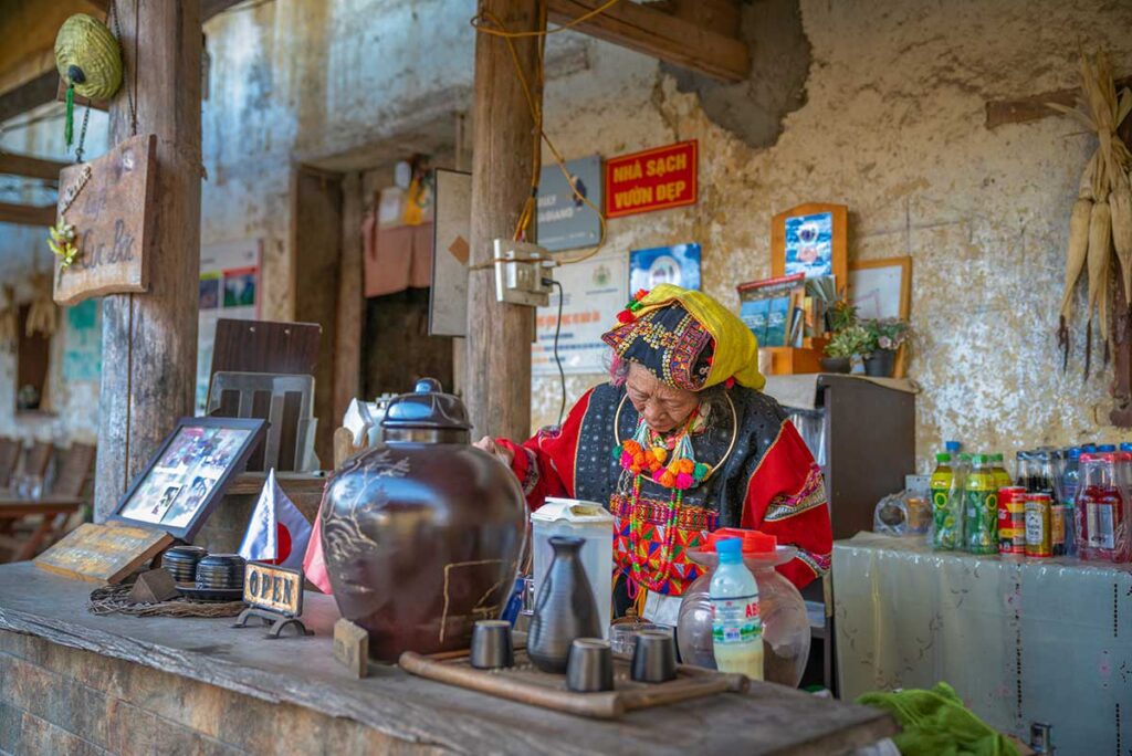 Elderly ethnic minority woman preparing tea inside a traditional home in Ha Giang, surrounded by local household items