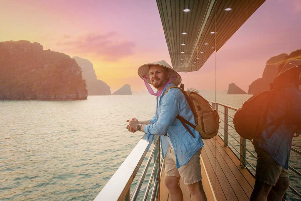 Traveler standing on a Halong Bay cruise deck at sunset, overlooking limestone karsts and calm sea in warm evening light