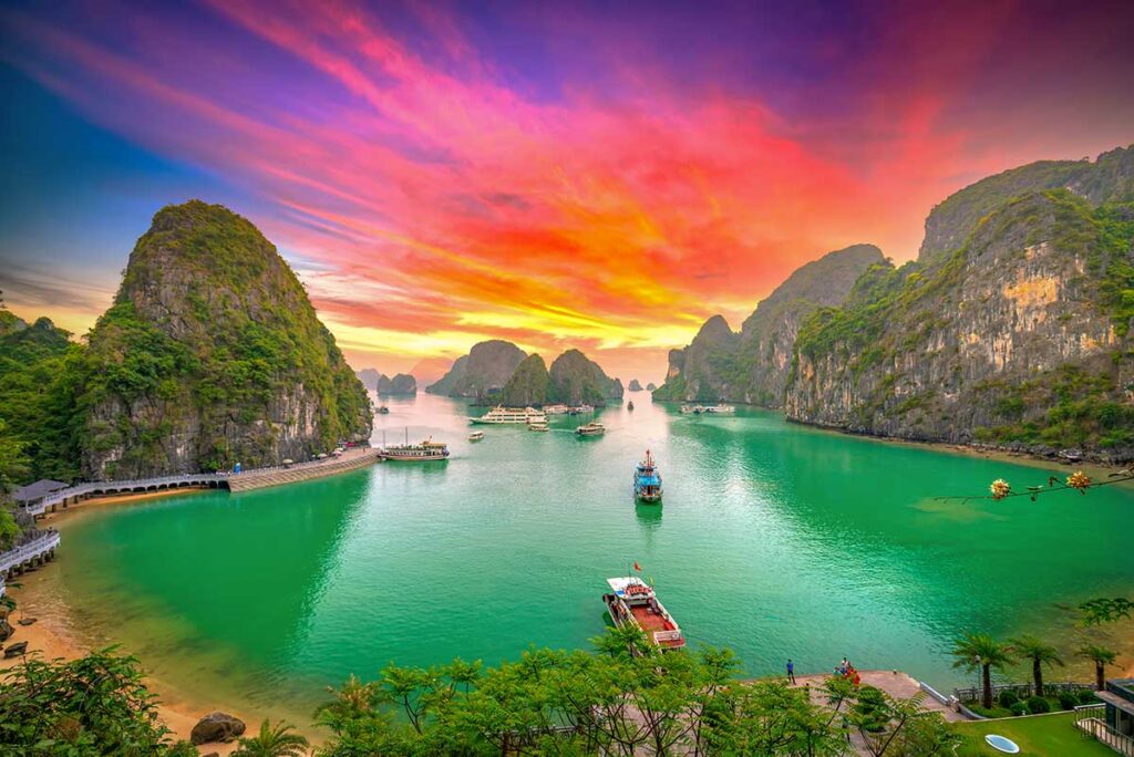 Halong Bay at sunset with colorful sky, limestone karsts, and boats on emerald water viewed from an elevated viewpoint