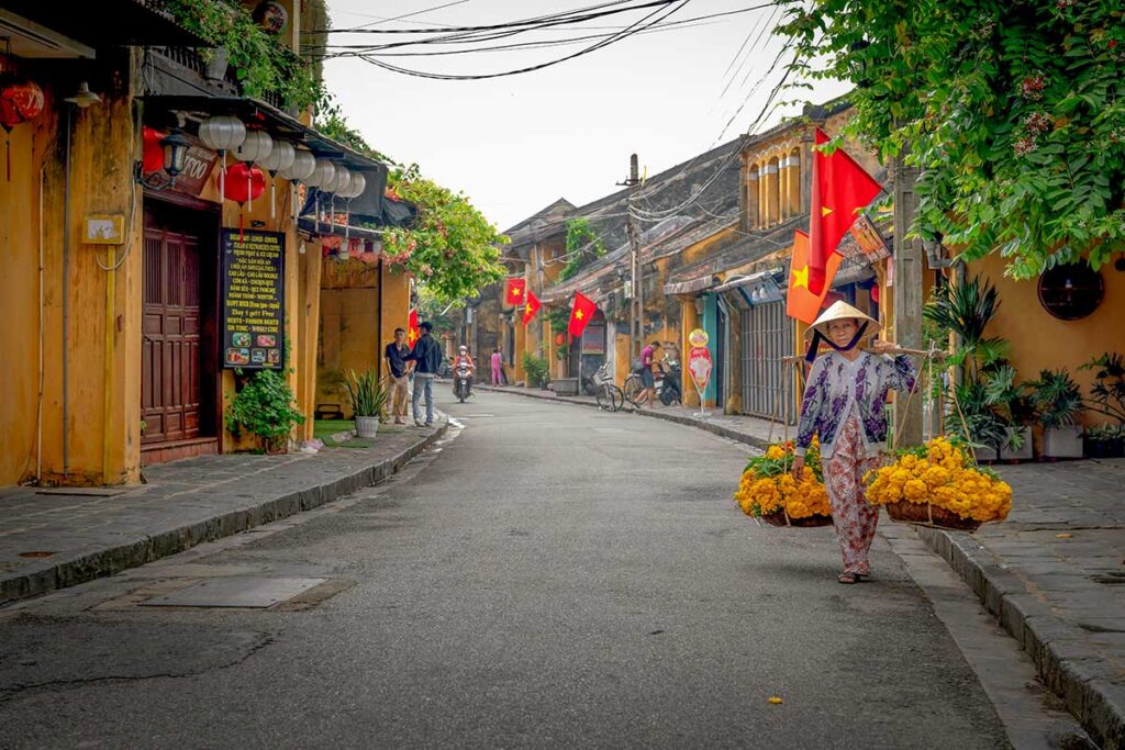 Local woman carrying flowers through a quiet street in Hoi An Ancient Town, with yellow houses and Vietnamese flags lining the road