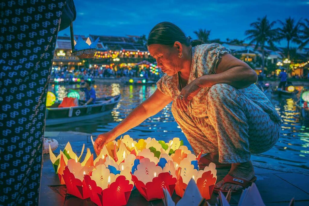 Local woman arranging colorful lanterns along the river in Hoi An at night, with glowing lights and boats in the background