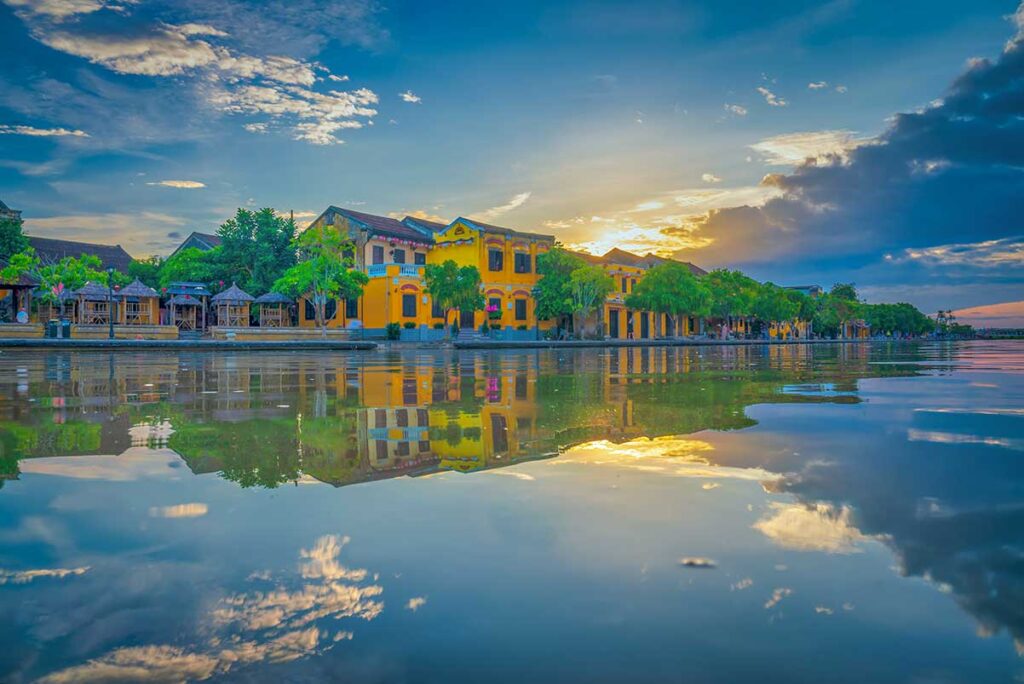 Hoi An Old Town along the river at sunset, with yellow colonial buildings reflected in calm water and a colorful sky above