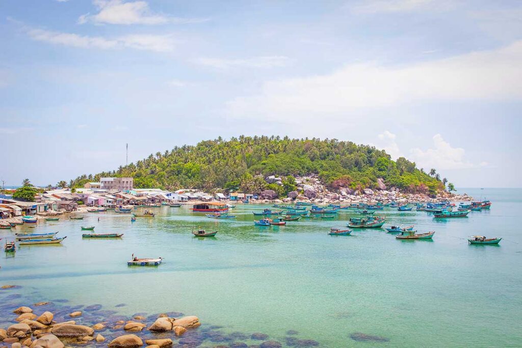 Fishing boats anchored off the coast of Hon Son Island, Tien Giang Province, with a backdrop of palm-covered hills and rocky shoreline.