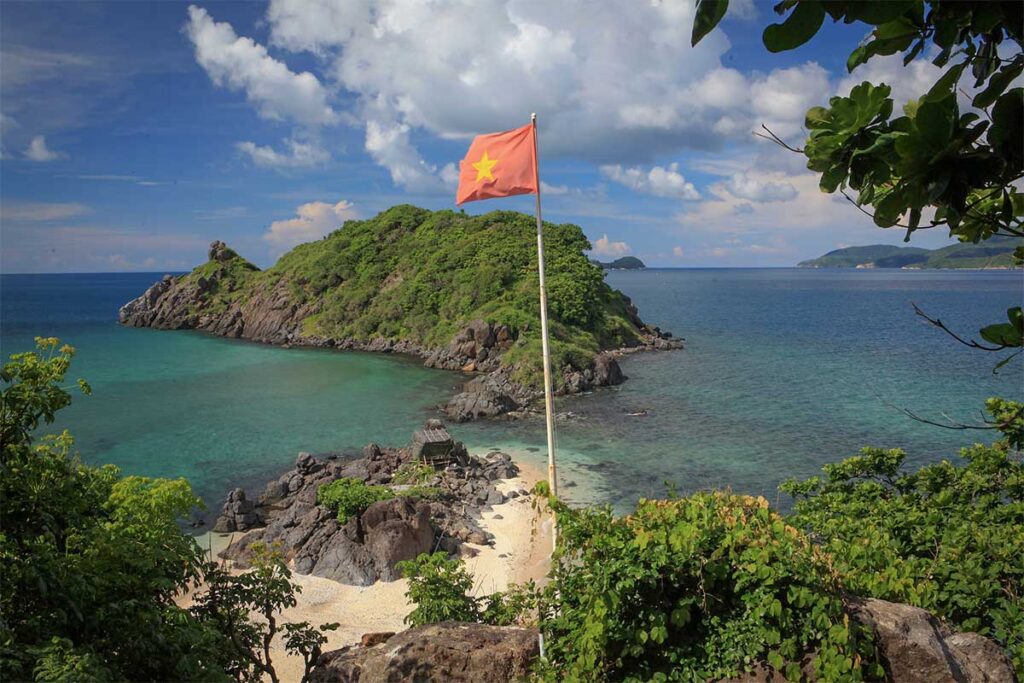 View of Hon Tre Lon Island in Con Dao with a Vietnamese flag overlooking a small beach and rocky coastline surrounded by clear blue sea.