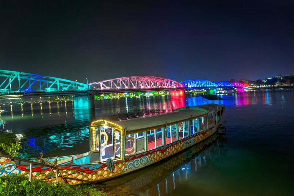 Colorful lit bridge over the Perfume River in Hue at night, with reflections on the water and a boat docked in the foreground