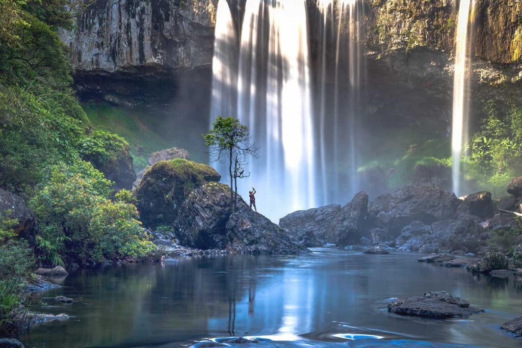 A traveler gazing up at the powerful K50 Waterfall, surrounded by mist and dense jungle in Gia Lai Province.