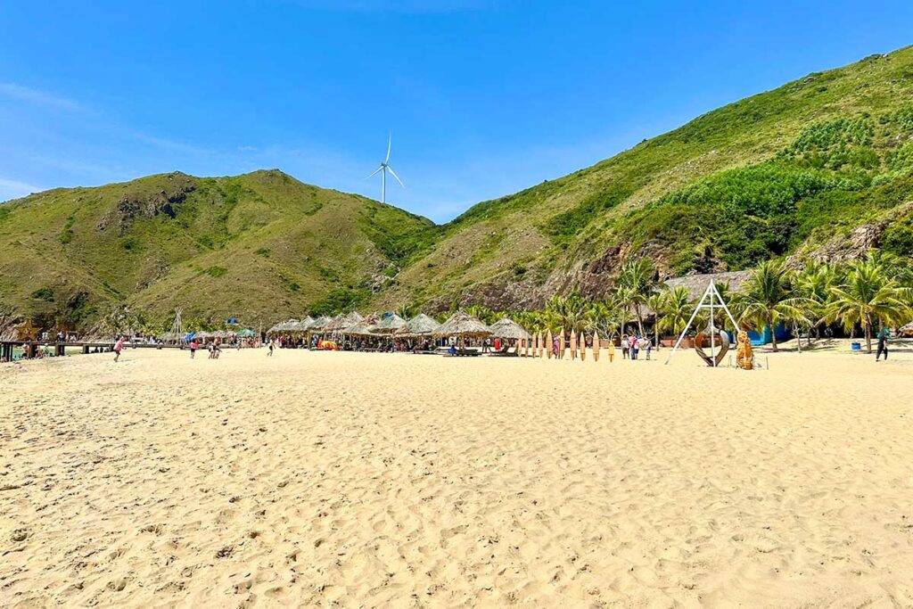 Sandy stretch of Ky Co Beach in Quy Nhon, Vietnam, with sunshades, tourists, palm trees, and a wind turbine overlooking from the hills.