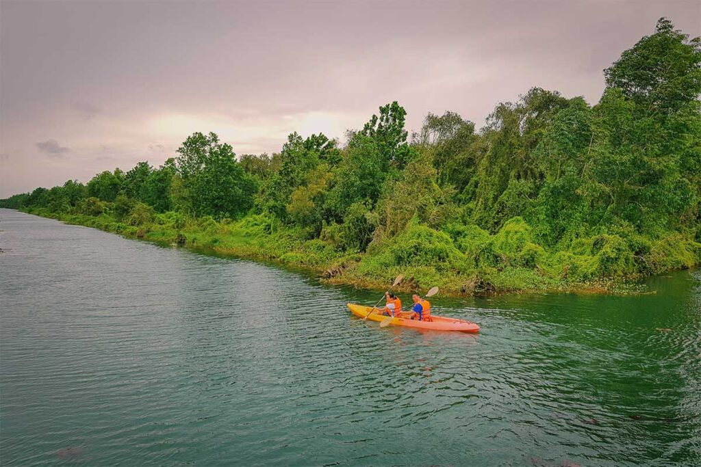 Two travelers kayaking along a forested canal in Long An's Endless Field eco-tourism area, surrounded by dense green foliage and calm waters.