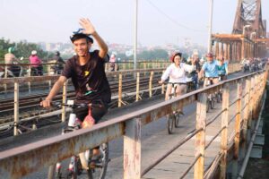 Local biking guide waving while leading tourists over Long Bien Bridge on a Hanoi bike tour