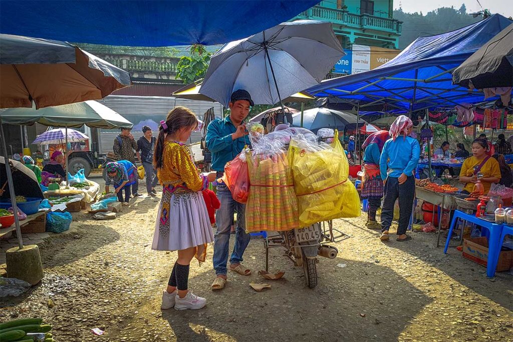 A young minority girl dressed in bright and gold colors is buying a snack from a minority man on a motorbike at the Lung Phin Market