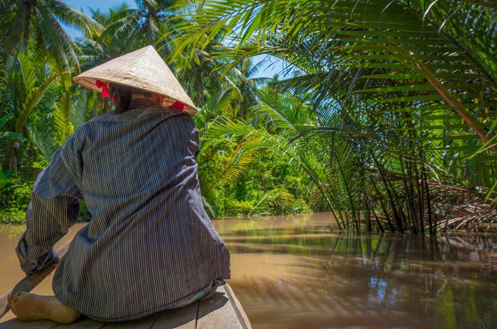 Person wearing a conical hat sitting in a small boat navigating narrow waterways surrounded by dense palm trees in the Mekong Delta