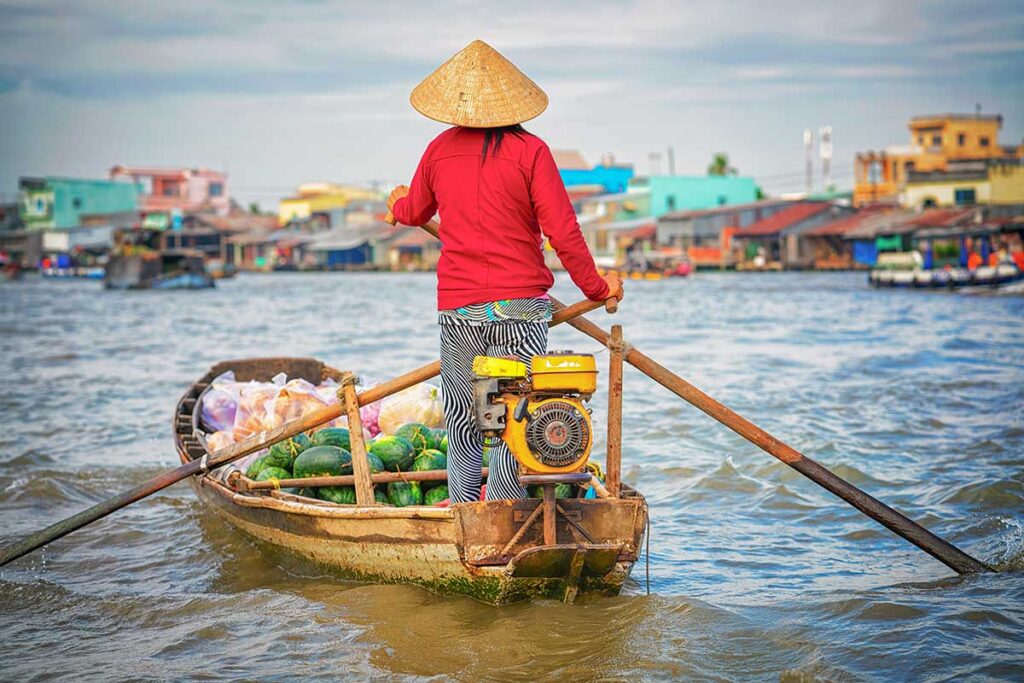 Local woman steering a small boat filled with fruit in the Mekong Delta, with floating market activity and riverside houses in the background