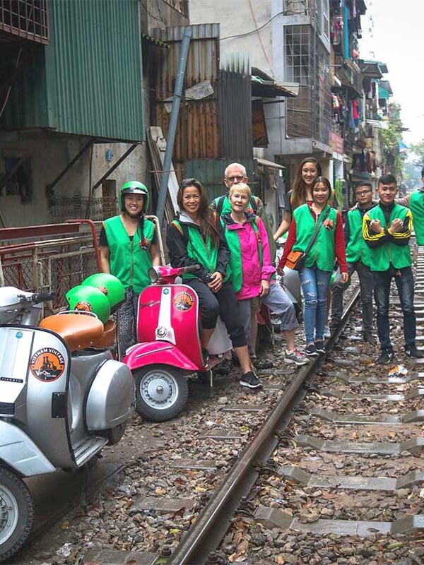 Group of travelers on Hanoi Train Street with Vespa motorbikes parked nearby during a motorbike tour