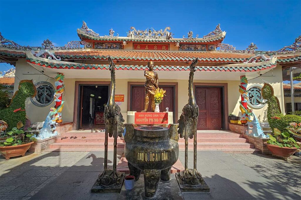 Exterior of Nguyen Trung Truc Temple in Rach Gia, Vietnam, with a statue of the national hero flanked by brass crane statues in front of the main altar.